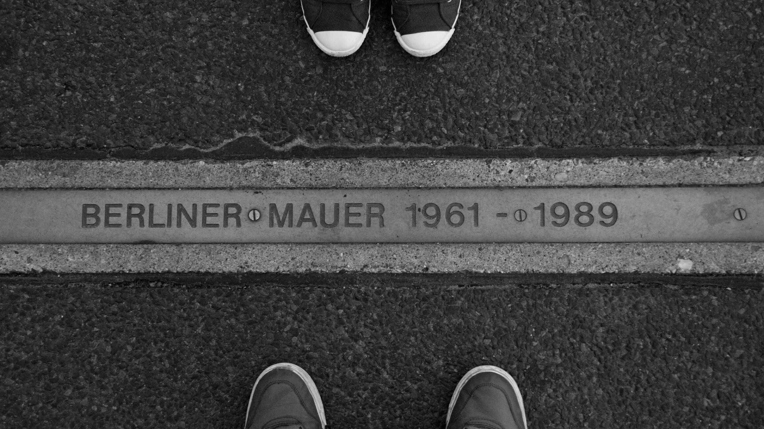 Schwarz-Weiß-Foto mit der Bodenmarkierung „Berliner Mauer 1961–1989“. Auf beiden Seiten stehen Menschen, von denen nur die Schuhe zu sehen sind.