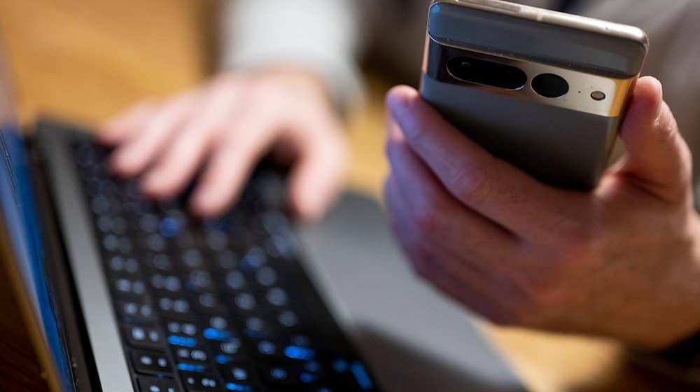 Ein Mann sitzt am Laptop und hält ein Smartphone mit in der Hand. / Foto: Monika Skolimowska/dpa