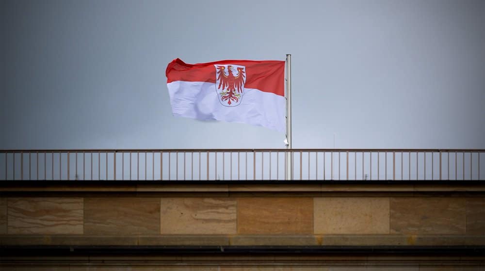 Die Flagge des Landes Brandenburg mit dem roten Adler weht auf dem Landtag vor grauem Himmel. / Foto: Soeren Stache/dpa