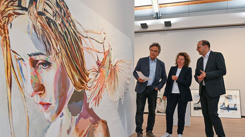 Frank Mangelsdorf (l), Jury-Vorsitzender, Heike Kramer, Geschäftsführerin der Stiftung Schloss Neuhardenberg und Tobias Dünow (SPD), Kulturstaatssekretär, stehen neben einem Gemälde von Cornelia Schleime. / Foto: Patrick Pleul/dpa