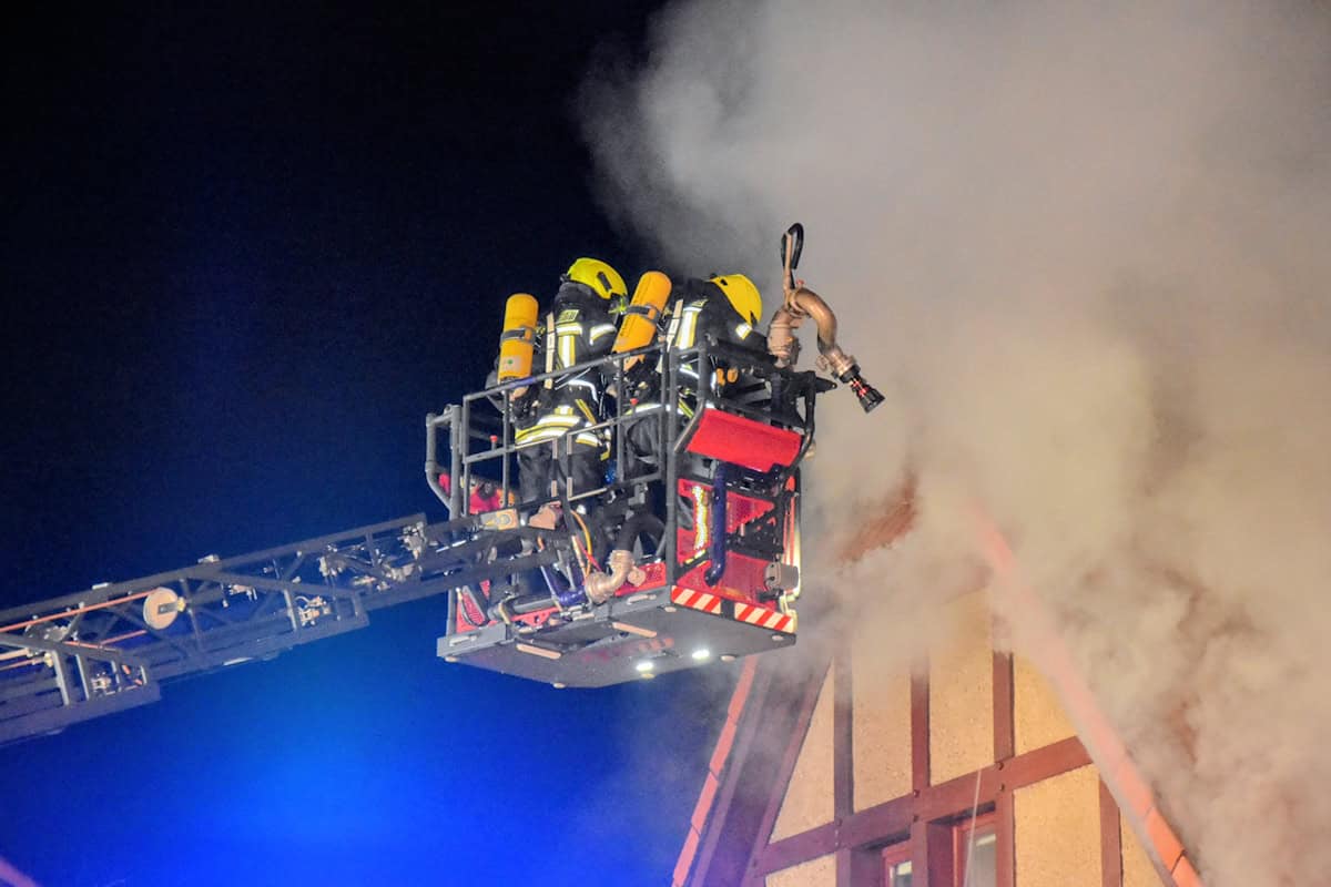 Mehrere Feuerwehrkräfte in gelben Helmen und Atemschutzgeräten auf einer Drehleiter vor einem rauchenden Fachwerkhaus bei Nacht. Mehrere Feuerwehrkräfte in gelben Helmen und Atemschutzgeräten auf einer Drehleiter vor einem rauchenden Fachwerkhaus bei Nacht.