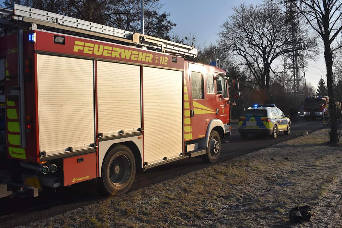 Rotes Feuerwehrfahrzeug steht auf einer Straße, dahinter zwei Einsatzfahrzeuge mit Blaulicht, umgeben von kahlen Bäumen
