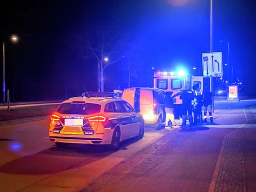 Polizeiauto und Einsatzfahrzeug mit Blaulicht an einer Straße bei Nacht, mehrere Personen stehen neben einem weißen Fahrzeug