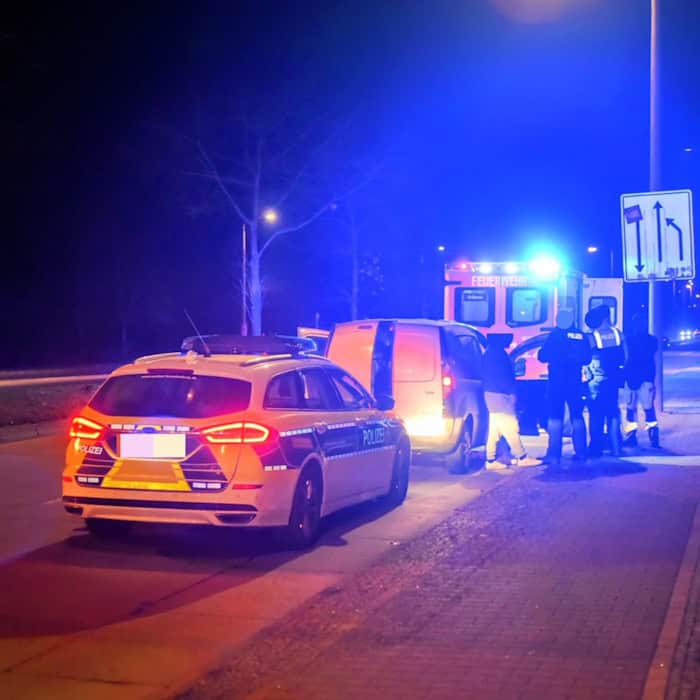 Polizeiauto und Einsatzfahrzeug mit Blaulicht an einer Straße bei Nacht, mehrere Personen stehen neben einem weißen Fahrzeug