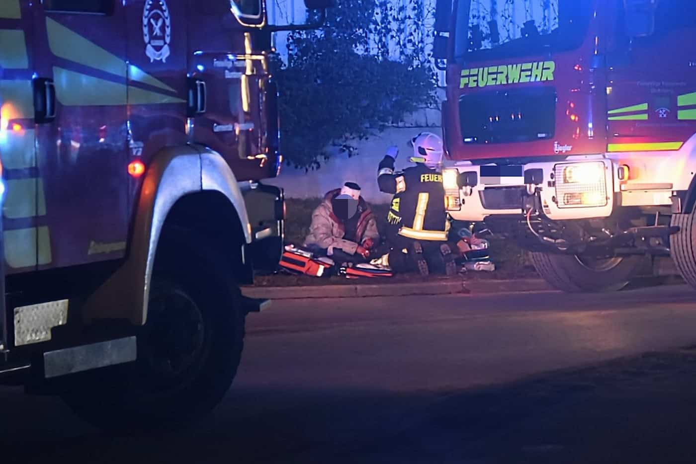 Zwei Feuerwehrleute in Schutzkleidung versorgen eine verletzte Person auf dem Boden sitzend zwischen zwei Feuerwehrfahrzeugen bei Nacht