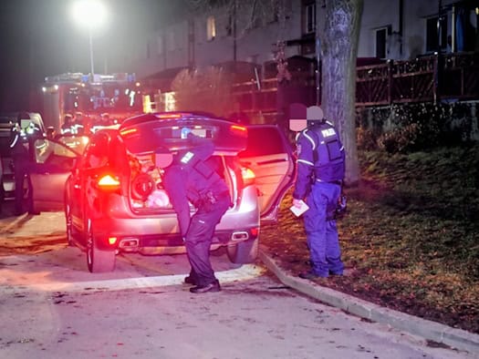 Polizisten durchsuchen nachts ein geöffnetes Fahrzeug auf einer Straße, im Hintergrund sind weitere Einsatzfahrzeuge mit eingeschalteten Lichtern zu sehen.