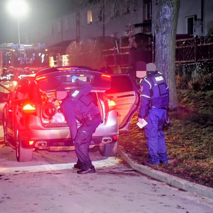 Polizisten durchsuchen nachts ein geöffnetes Fahrzeug auf einer Straße, im Hintergrund sind weitere Einsatzfahrzeuge mit eingeschalteten Lichtern zu sehen.