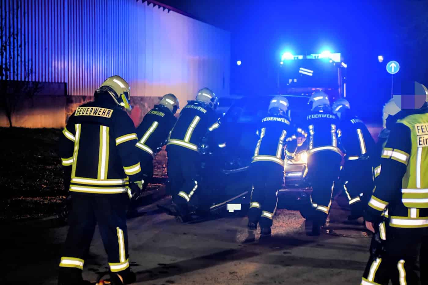 Mehrere Feuerwehrleute in Schutzkleidung schieben nachts ein Fahrzeug an, im Hintergrund leuchtet ein Einsatzfahrzeug mit Blaulicht