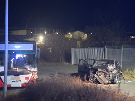 Beschädigter Bus und stark zerstörtes Auto mit geöffneter Fahrertür auf einer Straße bei Nacht.