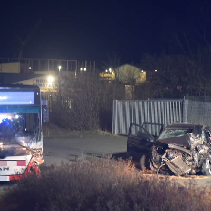 Beschädigter Bus und stark zerstörtes Auto mit geöffneter Fahrertür auf einer Straße bei Nacht.