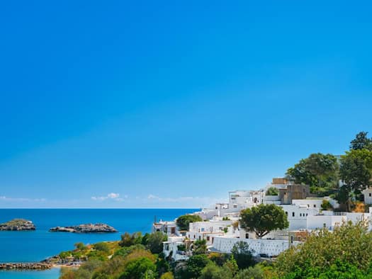Blick auf die Bucht und die schneeweißen Häuser der antiken Stadt Lindos auf der griechischen Insel Rhodos mit der Ägäis und den Inseln des Dodekanes-Archipels im Hintergrund.