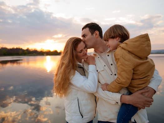 Eine Familie (Mann, Frau, Kind) steht am Ufer eines Sees bei Sonnenuntergang. Das Kind wird auf dem Arm getragen, während sich die Eltern umarmen.