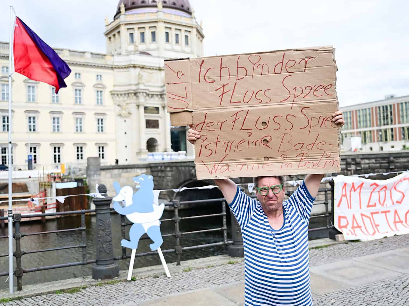 Ein Mann steht während der Schwimm-Demonstration gegen das Badeverbot in der Spree mit einem Schild am Ufer. / Foto: Sebastian Christoph Gollnow/dpa