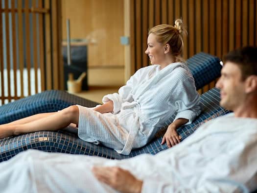 Zwei Personen in weißen Bademänteln entspannen auf blauen Liegen in einem Wellness-Bereich.