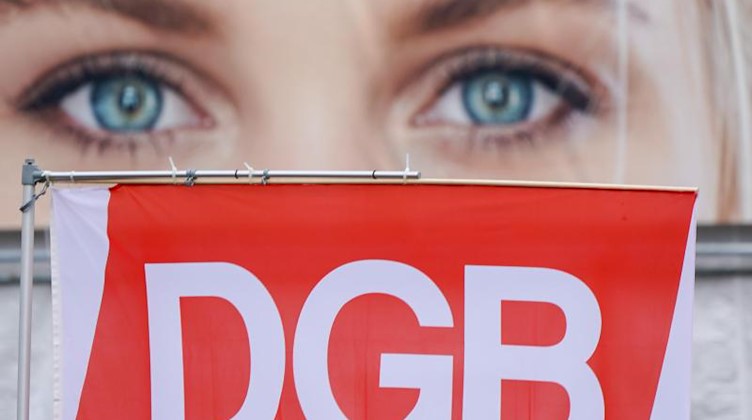 Ein Augenpaar von einem Werbeplakat schaut über einer Fahne des DGB. Foto: Peter Endig/dpa
