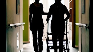Eine Pflegekraft begleitet die Bewohnerin eines Altenheims mit Rollator beim Gang durch den Flur. Foto: Oliver Berg/dpa/Symbolbild