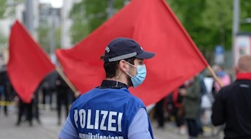Ein Polizist mit Maske und einer Weste "Polizei Kommunikationsteam" steht am Rande einer linken Demonstration zum 1. Mai. Foto: Sebastian Willnow/dpa-Zentralbild/dpa