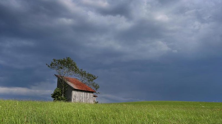 Dunkle Regenwolken ziehen über eine Holzhütte und eine Wiese die noch von der Sonne beschienen werden. Foto: Karl-Josef Hildenbrand/dpa/Archivbild