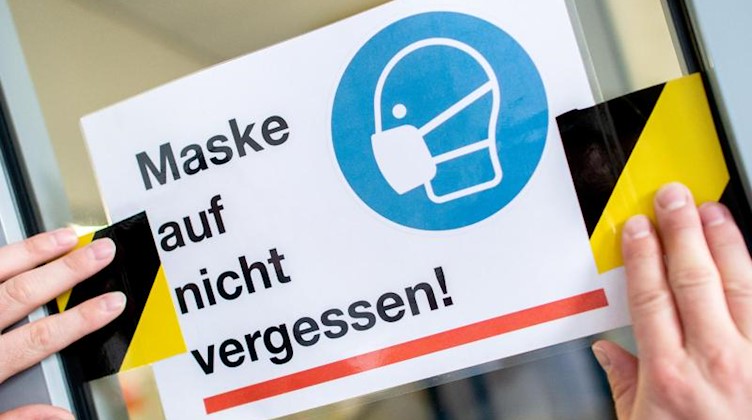 Ein Schild mit der Aufschrift «Maske auf nicht vergessen!» wird an eine Glasscheibe geklebt. Foto: Hauke-Christian Dittrich/dpa/symbol