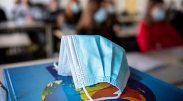 Ein Mund-Nasen-Schutz liegt während des Unterrichts auf einem Atlas. Foto: Matthias Balk/dpa/Illustration