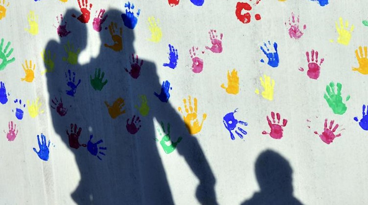 Ein Mann und zwei Kinder werfen Schatten auf die mit Handabdrücken bemalte Wand einer Kindertagesstätte. Foto: Peter Kneffel/dpa/Symbolbild