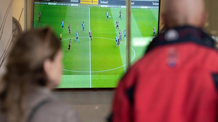 Zwei Menschen verfolgen außerhalb einer Sportsbar ein Bundesliga-Spiel auf einem Fernseher. Foto: Silas Stein/dpa/Symbolbild