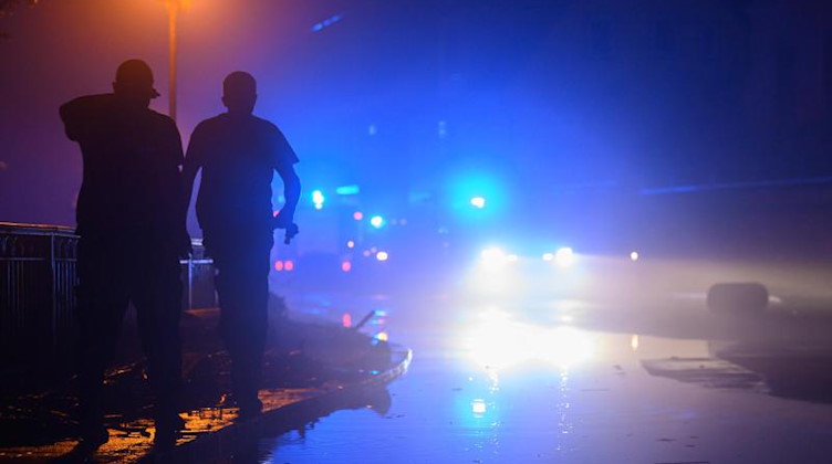 Zwei Männer laufen am Rande einer von Wasser der Kirnitzsch überfluteten Straße entlang während im Hintergrund ein Feuerwehreinsatz läuft. Foto: Robert Michael/dpa-Zentralbild/dpa