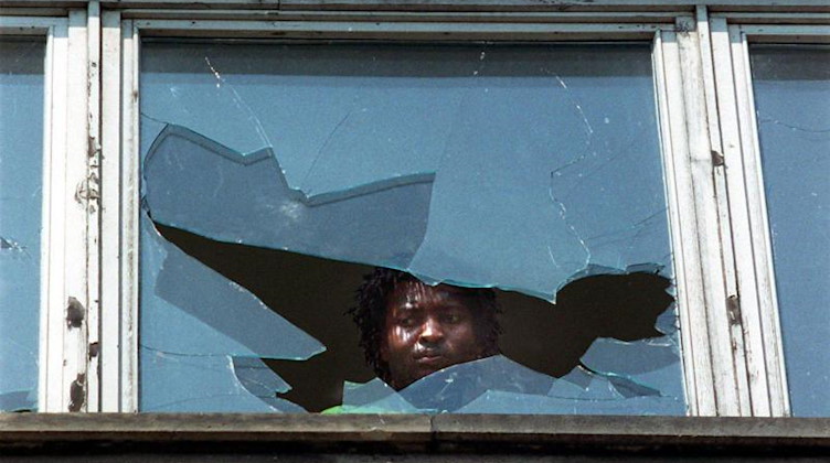 Ein Mann blickt am Tag nach den ausländerfeindlichen Übergriffen durch die eingeworfene Scheibe eines Wohnheims. Foto: Rainer Weisflog/dpa-Zentralbild/dpa/Archivbild