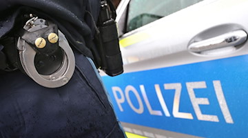 Ein Polizist mit Handschellen und einer Pistole am Gürtel steht vor einem Streifenwagen. / Foto: Karl-Josef Hildenbrand/dpa