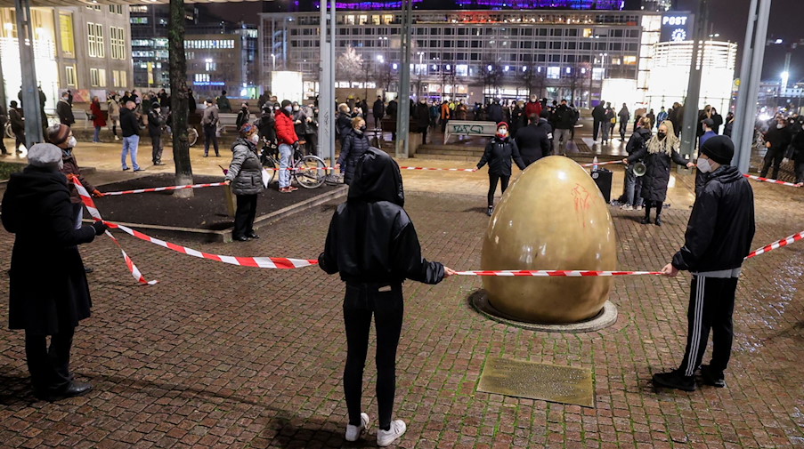 Demonstranten gegen die Anti-Corona-Proteste halten ein rot-weisses Absperrband um die «Demokratieglocke» in Leipzig. / Foto: Jan Woitas/dpa