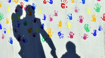 Ein Mann mit einem Kind auf dem Arm und einem an der Hand wirft einen Schatten auf eine Wand. / Foto: Peter Kneffel/dpa/Archivbild