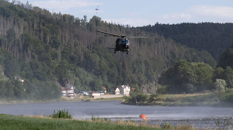 Ein Hubschrauber der Bundeswehr fliegt mit einem Löschwasser-Außenlastbehälter über der Elbe. / Foto: Sebastian Kahnert/dpa