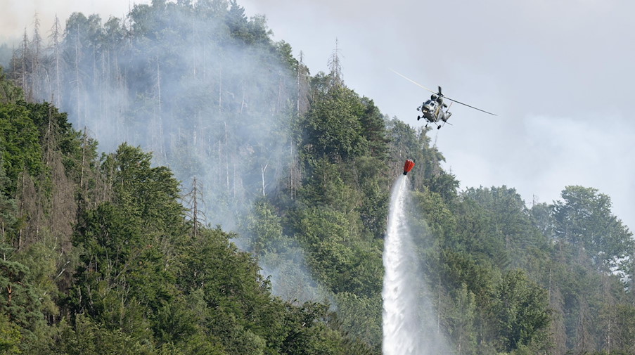 Ein Löschhubschrauber fliegt mit einem Löschwasser-Außenlastbehälter über einen Brand im Nationalpark. / Foto: Sebastian Kahnert/dpa