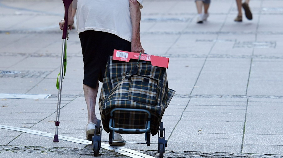 Eine alte Frau mit einer Krücke und Einkaufstrolley geht durch eine Fußgängerstraße. / Foto: Jens Kalaene/dpa/Archivbild