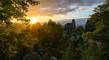 Die Sonnen geht am Morgen über dem Nationalpark Sächsische Schweiz neben der Basteibrücke auf. / Foto: Robert Michael/dpa/Archivbild