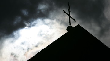Im Gegenlicht und vor wolkenverhangenem Himmel ist die Kirchturmspitze des Doms mit Kreuz zu sehen. / Foto: Friso Gentsch/dpa/Symbolbild