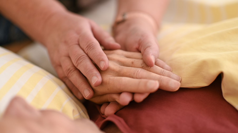 Eine Hospizmitarbeiterin hält die Hand eines todkranken Menschen, der in einem Hospiz im Bett liegt. / Foto: Felix Kästle/dpa/Symbolbild