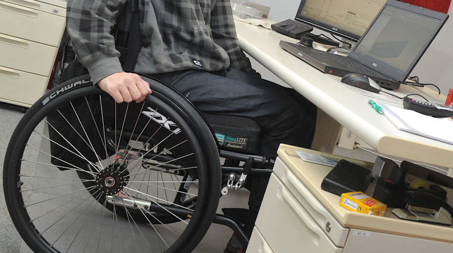 Ein Mann sitzt in einem Rollstuhl an seinem Arbeitsplatz am Schreibtisch. / Foto: Stefan Puchner/dpa
