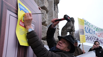 Ein Umweltaktivist nagelt ein Plakat mit den Begriffen „Jobwunder“ und „Killerjob“ an eine symbolische Tür vor dem Neuen Rathaus. / Foto: Sebastian Willnow/dpa