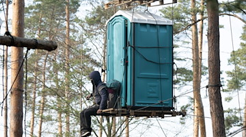 Ein Aktivist der Initiative «Heibo bleibt» sitzt in einem besetzten Waldgebiet an einem mit Seilen befestigten Dixi-Klo. / Foto: Sebastian Kahnert/dpa/Archivbild