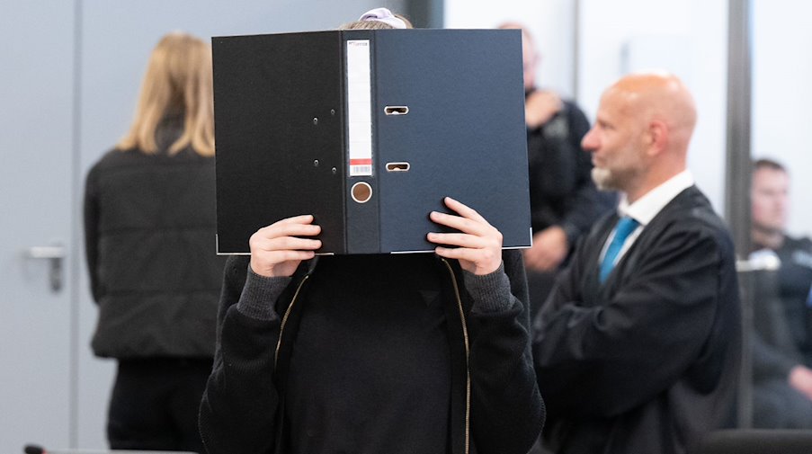 Die Angeklagte Lina E. steht bei der Fortsetzung des Prozesses im Oberlandesgericht (OLG) Dresden im Verhandlungssaal und hält einen Aktenordner vor ihr Gesicht. / Foto: Sebastian Kahnert/dpa