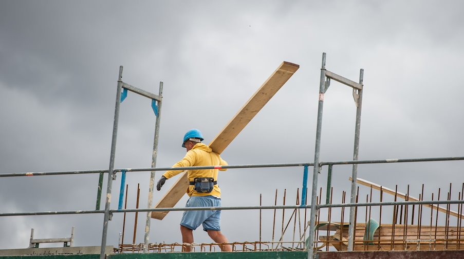 Ein Bauarbeiter trägt auf einer Baustelle auf einem Hausdach ein Brett. / Foto: Christophe Gateau/dpa