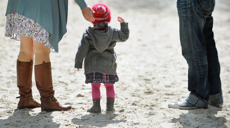 A child stands between his parents on a playground. / Photo: picture alliance / Andreas Gebert/dpa/iconic image