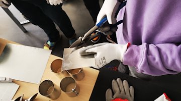 Vista desde arriba de un alumno cortando una chapa. Manos con guantes de trabajo cortan una pieza de chapa con cizallas para metal. Al fondo, una mesa con piezas cortadas y dobladas en cilindros.