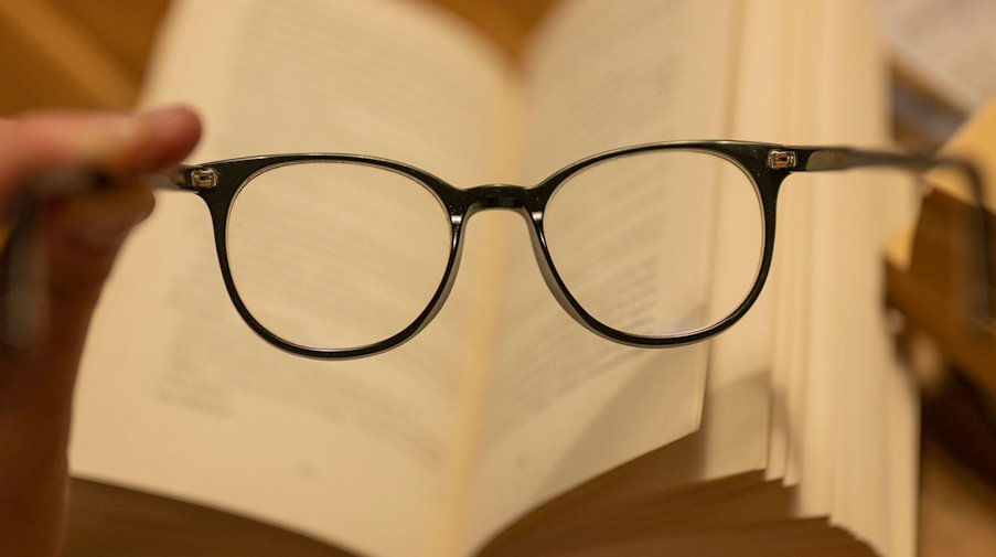 Un hombre sostiene unas gafas de lectura delante de un libro de papel abierto / Foto: Monika Skolimowska/dpa/Illustration