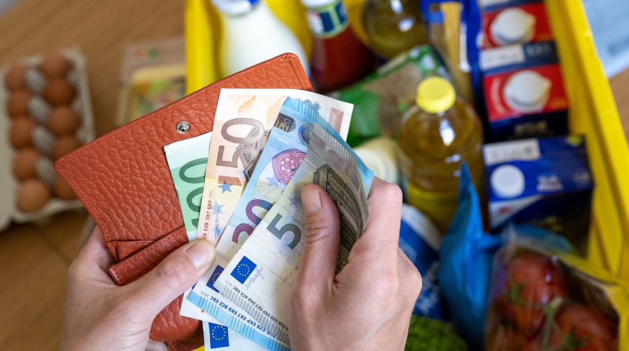 Una caja de la compra con comestibles sobre la mesa de una cocina mientras una mujer sostiene billetes de euro en las manos. / Foto: Hendrik Schmidt/dpa