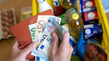 Una caja de la compra con comestibles sobre la mesa de una cocina mientras una mujer sostiene billetes de euro en las manos. / Foto: Hendrik Schmidt/dpa