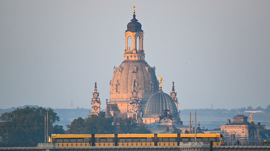 Eine Straßenbahn der Dresdner Verkehrsbetriebe (DVB) fährt am Morgen vor der Altstadtkulisse mit der Frauenkirche (l) und der Kuppel der Kunstakedmie mit dem Engel «Fama» über die Albertbrücke. / Foto: Robert Michael/dpa