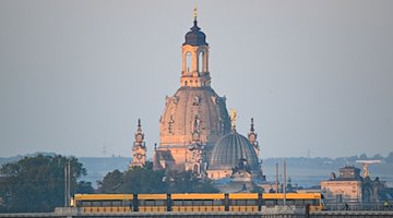 Eine Straßenbahn der Dresdner Verkehrsbetriebe (DVB) fährt am Morgen vor der Altstadtkulisse mit der Frauenkirche (l) und der Kuppel der Kunstakedmie mit dem Engel «Fama» über die Albertbrücke. / Foto: Robert Michael/dpa