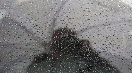 Regentropfen haften an einer Fensterscheibe eines Fahrzeugs, in der sich eine Person mit Regenschirm spiegelt. / Foto: Matthias Bein/dpa